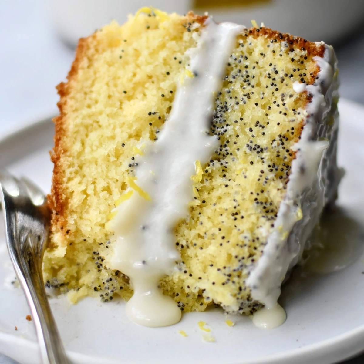 A moist slice of Lemon Poppy Seed Cake with Icing is plated, showcasing golden crumb and speckled seeds.