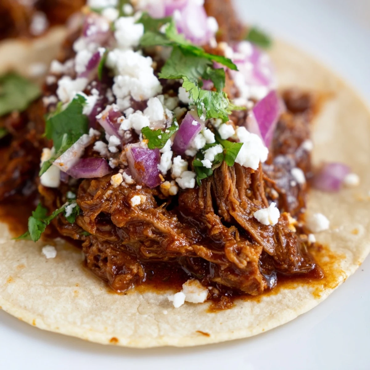 Slow cooker shredded beef barbacoa tacos topped with cilantro, onion, and crumbled queso fresco on warm corn tortillas.