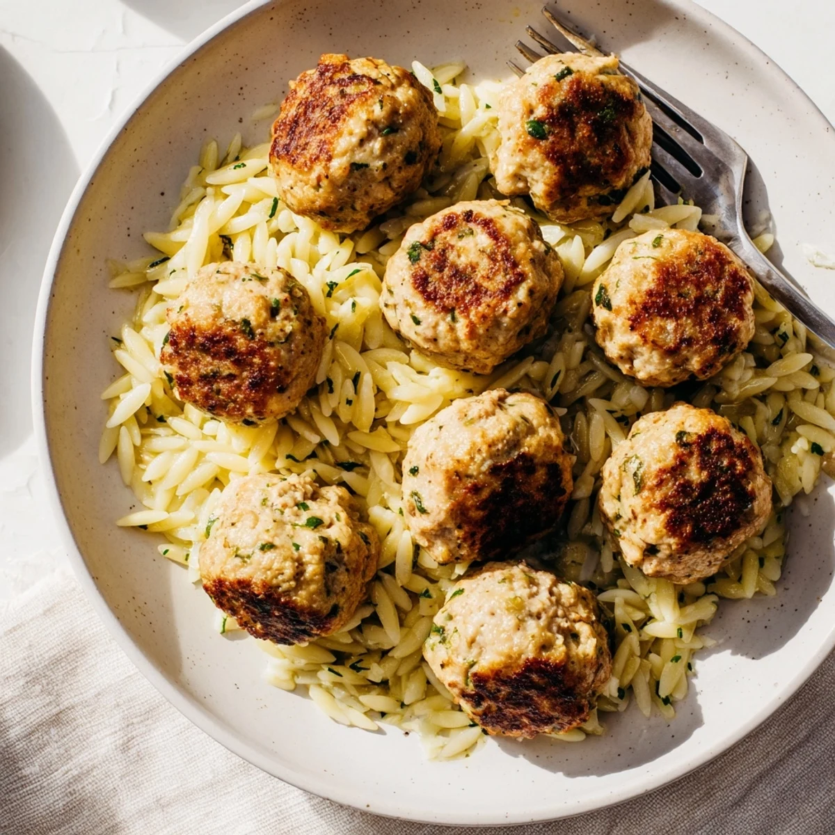 Golden brown chicken meatballs nestled over bright lemon orzo with fresh herbs in a white bowl