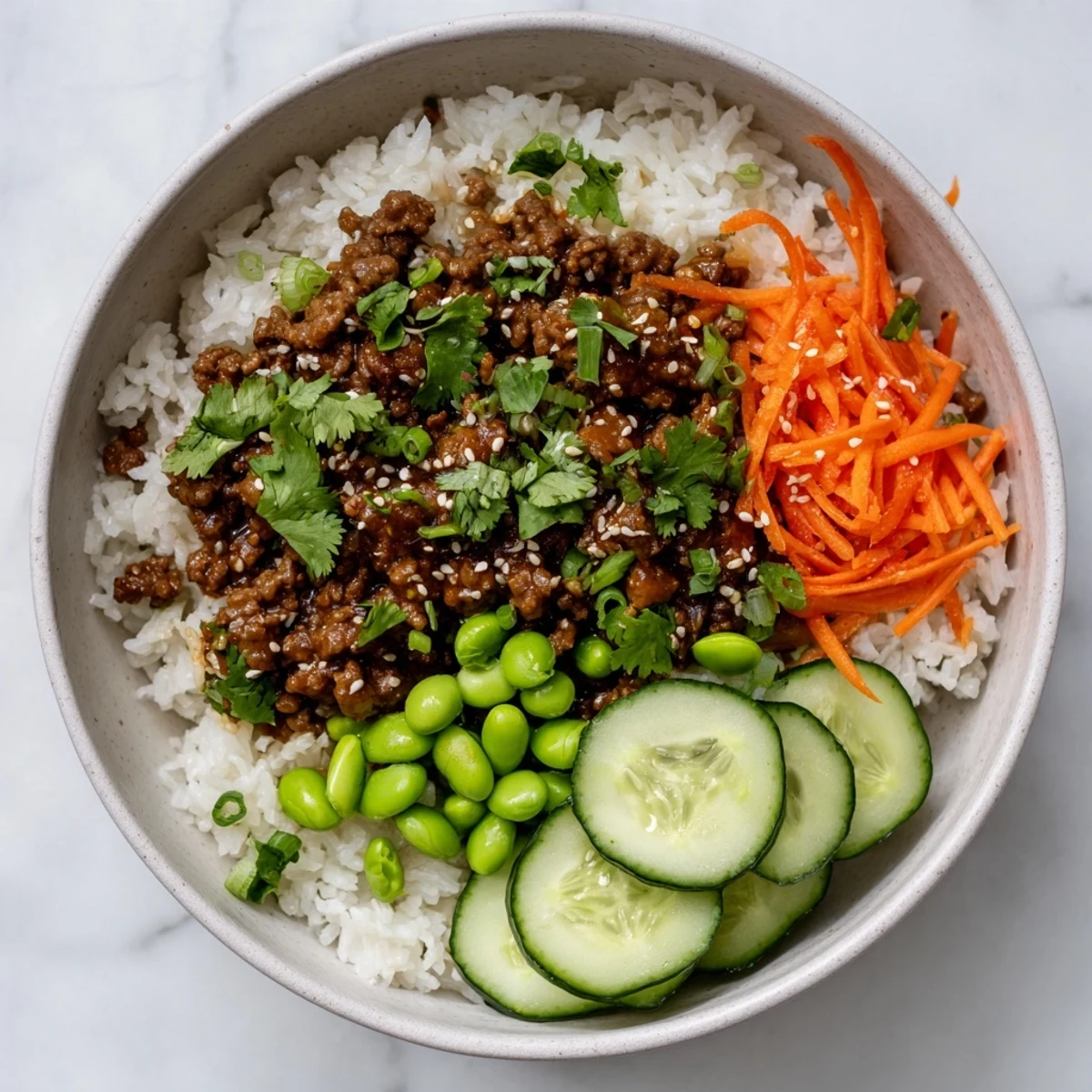 Glossy ground beef glazed in spicy hot honey sauce arranged in a bowl with edamame, carrots, and cucumber toppings.