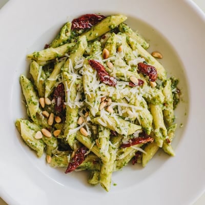 Sun-drenched Italian-inspired Green Pesto Pasta with Sun-Dried Tomatoes on a rustic plate, garnished with pine nuts and a light drizzle of olive oil.